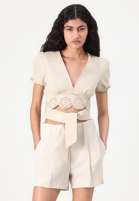 Top beige court avec manches bouffantes, décolleté en V, et motif circulaire découpé décoratif à l'ourlet. Associé à un short taille haute et une ceinture nouée.