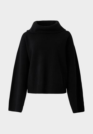 Woman wearing loose black pants and a black sweater holding a large black leather bag on her shoulder against a plain white background.