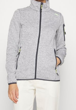 Persoon die een lichtgrijs, gestructureerd jack met rits draagt, met zwarte afwerking, ritszakken en lichtgekleurde broek, met één hand in een zak.