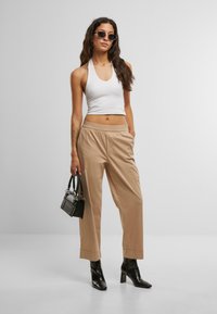 Pantaloni beige a gamba larga con cintura elastica, abbinati a un top bianco aderente senza maniche. Gli accessori includono una piccola borsa nera e occhiali da sole rotondi.