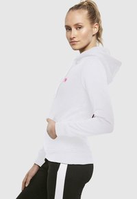 Hoodie bianco realizzato in morbido tessuto con tasca a marsupio. Presenta un logo rosa sul petto e polsini a coste, abbinato a pantaloni neri.