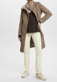 Femme portant un manteau en laine marron, un pull marron foncé par-dessus une chemise rayée, un pantalon crème et des baskets crème, debout avec une main sur la hanche.