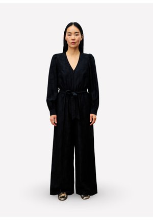 Femme aux longs cheveux noirs portant une combinaison noire à manches longues avec une ceinture à la taille et des ballerines métalliques, se tenant sur un fond blanc.