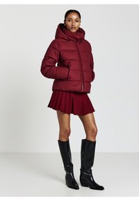 Donna che indossa un giubbotto imbottito bordeaux, una gonna plissettata abbinata e stivali neri al ginocchio, in piedi con le mani nelle tasche contro uno sfondo neutro.