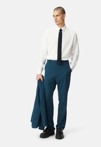 Witte overhemd, marineblauwe stropdas met stippen, teal broek en donkere schoenen. Model houdt een teal blazer vast en staat met een neutrale achtergrond.