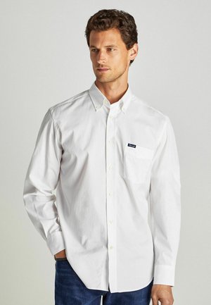 Camicia bianca a maniche lunghe con bottoni, dotata di tasca sul petto e realizzata in morbido cotone, abbinata a jeans blu scuro. Logo discreto sulla tasca.