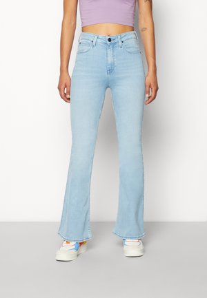 Femme portant un jean flare taille haute bleu clair, un top court lilas et des baskets blanches avec des détails multicolores, debout devant un fond blanc.