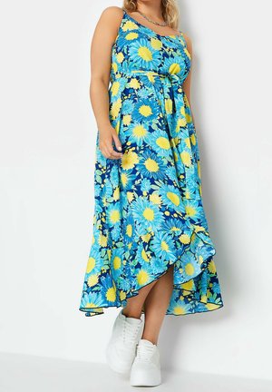 Donna che indossa un vestito midi senza maniche azzurro e giallo con motivo floreale e cintura in vita, abbinato a sneakers bianche, in piedi davanti a uno sfondo semplice.