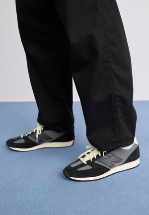 Black sneakers with grey accents, suede and mesh material, featuring white soles and light yellow laces. Rounded toe and low profile design.