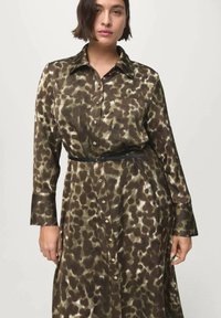 Žena v dlouhých rukávech, s hnědo-beige šaty s potiskem zvířat, zapnutými na knoflíky a s černým opaskem na světlém jednobarevném pozadí.