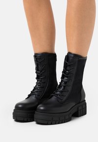 Black leather lace-up ankle boots with chunky soles worn on bare legs against white background.