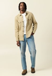 Beige knäppt skjorta med två bröstfickor, bärs över en vit skjorta; kombineras med ljusblå jeans och bruna ankelstövlar.