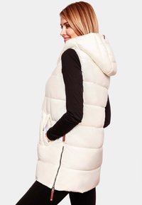 Gilet matelassé blanc avec capuche, doté d'une fermeture éclair et de poches latérales. Texture lisse avec un design matelassé, porté par-dessus un haut noir.
