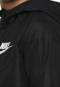 Mustan kevyt takin huppu, teksturoitu kangas ja selkeä valkoinen Nike-logo rinnassa.