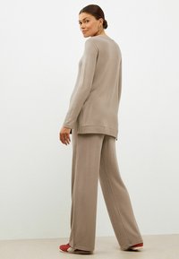 Hellbeige Strickpullover mit langen Ärmeln und seitlichen Schlitzen, kombiniert mit beigen weit geschnittenen Hosen. Das Model trägt rote Schuhe. Einfarbiger Hintergrund.