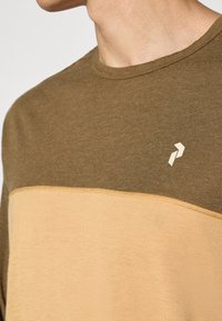 Mann som har på seg en t-skjorte med to farger, med olivengrønn øverste del, beige nederste del og en liten hvit geometrisk logo på brystet.