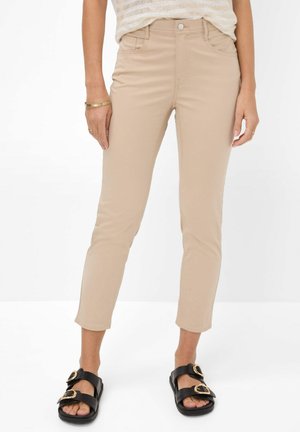 Beige, kurz geschnittene, eng anliegende Hose an einer Person, die schwarze Sandalen mit goldenen Schnallen und ein leicht gestreiftes Oberteil trägt.