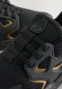 Baskets de sport noires avec accents dorés, dotées d'une tige en maille, de lacets texturés et d'une semelle en caoutchouc distinctive avec des détails mouchetés.