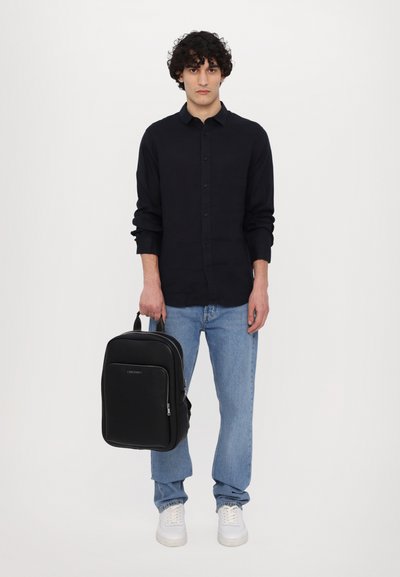 Sac à dos noir au design épuré, en matériau synthétique lisse, avec une poche zippée à l'avant et une poignée sur le dessus. Porté par une personne portant une chemise sombre et un jean.