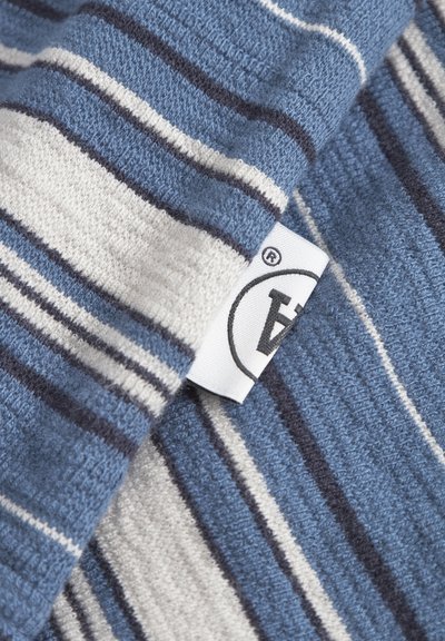 Tissu rayé aux nuances variées de bleu et de crème, avec une texture douce ; comprend une étiquette visible avec un logo et desinstructions d'entretien.