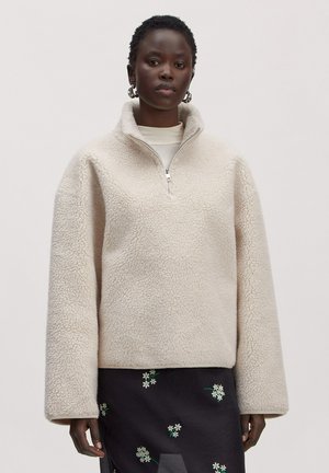 Femme portant un pull en polaire couleur crème avec une fermeture éclair au quart et une jupe noire avec de petites broderies florales blanches.