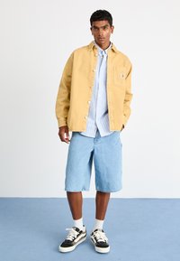 Gul skjorte med knappelukning, blå stribet skjorte under, lyseblå denim shorts, sort og hvidt sneakers, står på blå baggrund.