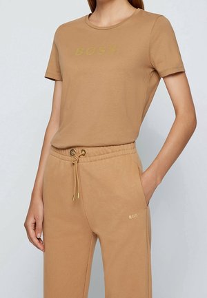 Vrouw draagt een beige T-shirt met korte mouwen en bijpassende broek met een gouden "BOSS"-logo op de borst en vlak bij de zak.