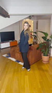 Mulher de cabelo comprido, vestindo um casaco azul e calças pretas, está numa sala de estar perto de um sofá castanho e uma grande planta em vaso.