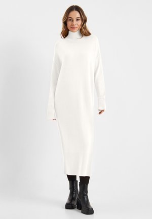 Witte, gebreide turtleneck jurk met lange mouwen en geribbelde manchetten, die tot halverwege de kuit reikt. Gepaard met zwarte chunky heeled boots.