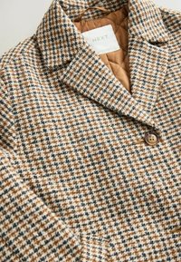 Brūns un balts houndstooth režģis ar quiltētu iekšējo oderi un pogu aizdari. Iedarbina izgriezta apkakle un teksturēta auduma.