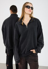 Čierny fleece pulóver s polovičným zipsom a nadmernou fit. Textúrovaná tkanina a dlhé rukávy. Model nosí tmavé nohavice.