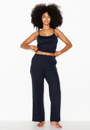 Donna in piedi a piedi nudi su sfondo bianco, indossa un top senza maniche blu navy e pantaloni ampi coordinati, mani sui fianchi.