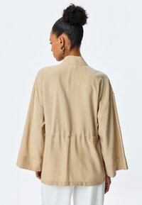 Beige leichte Jacke mit weit ausgestellten Ärmeln, lockerer Passform und geraffter Taille. Subtile Textur mit glatter Oberfläche und minimalistischem Design.
