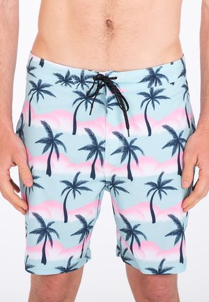 Man wearing light blue swim shorts with black palm tree and pink cloud patterns, black drawstring at waist.