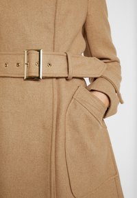 Cappotto di lana beige con un design strutturato, caratterizzato da una cintura larga, accessori in oro e tasche laterali. Texture morbida con un delicato motivo a intreccio.