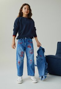 Fille portant un sweat-shirt bleu marine et un jean bleu brodé, tenant une veste en denim à côté d'un fauteuil bleu foncé sur fond blanc.