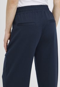 Personne portant un pantalon à taille élastique bleu marine avec deux poches passepoilées à l'arrière, debout avec la main gauche reposant près du côté.