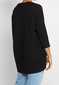 Czarny, oversize'owy sweter o fakturze typu waffle; posiada rękawy do połowy przedramienia i okrągły dekolt, zestawiony z jasnoniebieskimi dżinsami.