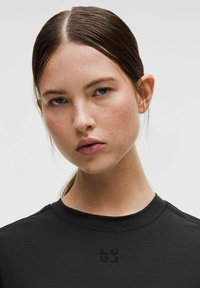 Fekete póló kerek nyakkivágással és rövid ujjakkal, középen egy diszkrét logóval. Sima textúra és testhezálló szabás.