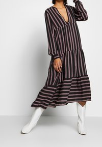 Černé šaty s vertikálními růžovými pruhy, výstřih do V, dlouhé rukávy a sukni ve vrstvách. Nosené s bílými kotníkovými botami. Hladká textilie.