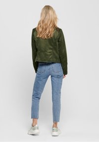 Giubbotto in ecopelle verde oliva con design fitted, tasche posteriori e dettagli delle cuciture, abbinato a jeans blu chiaro cropped e sneakers grigie.