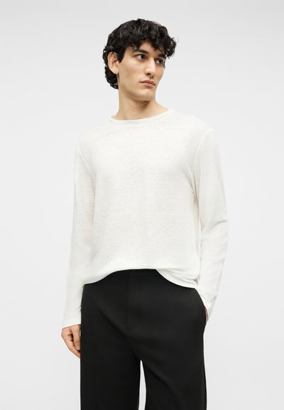 Νεαρός άνδρας με σγουρά μαλλιά που φοράει λευκό πουλόβερ μακρυμάνικο και μαύρο παντελόνι, κοιτάζει πλάγια με το ένα χέρι στην τσέπη.