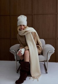 Beige suède jas, witte wollen muts, crème sjaal, bijpassende vingerloze handschoenen, donkerbruine kniehoge laarzen, zilveren stoel op een lichte vloer.