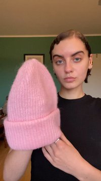 Ροζ μάλλινο σκουφάκι με ριγωτό μανίκι και αιχμηρή κορυφή, κρατιέται από ένα άτομο. Η υφή φαίνεται μαλακή και φουντωτή, κατάλληλη για κρύο καιρό.