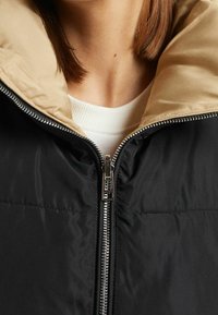 Black puffer jacket with beige collar, featuring a silver zipper and soft texture. Visible stitching and a high neckline.