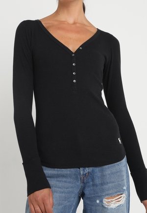 Woman wearing a fitted black long-sleeve V-neck top with buttons and distressed blue jeans with a rip on the thigh.