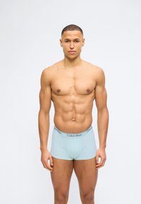 Μυώδης άνδρας που στέκεται με το πρόσωπο προς τα εμπρός φορώντας ανοιχτό μπλε εσώρουχα Calvin Klein σε λευκό απλό φόντο.