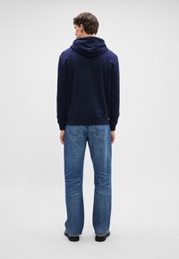 Hanorac cu glugă de culoare bleu marin, cu o textură netedă, asortat cu blugi drepți de culoare bleu deschis. Viziunea din spate arată o croială relaxată și manșete ribbate.