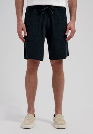Man met zwarte shorts met trekkoord en beige instapschoenen, staand tegen een effen lichte achtergrond.