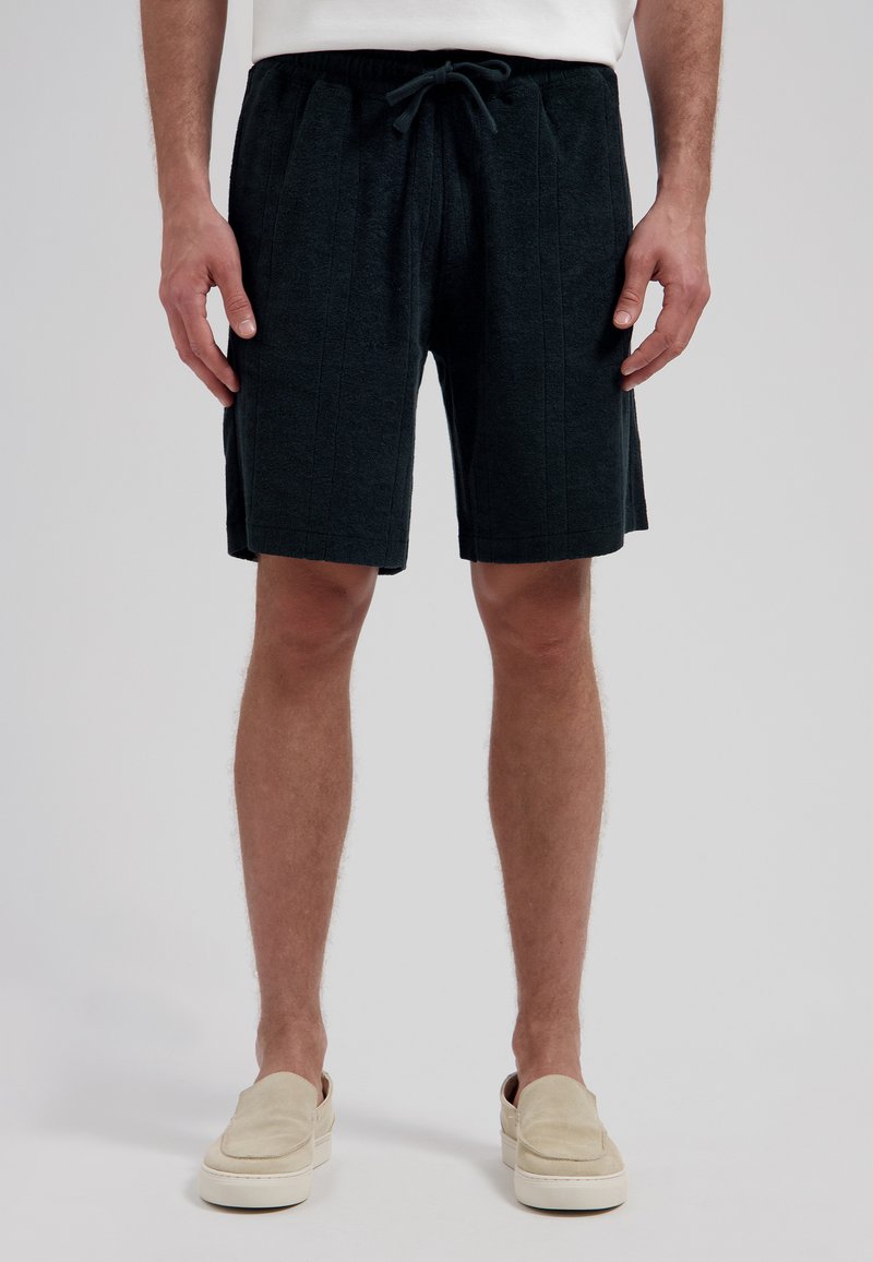 Homme portant un short noir à cordon de serrage et des chaussures beige à enfiler, debout devant un fond clair uni.